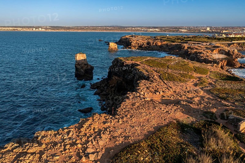 Bird's-eye view of ocean waves break on rocky shores pf Peniche, Portugal. Summer sunset haze, little foliage and rocky cliffs, peninsula and rocks, fishing town, horizon