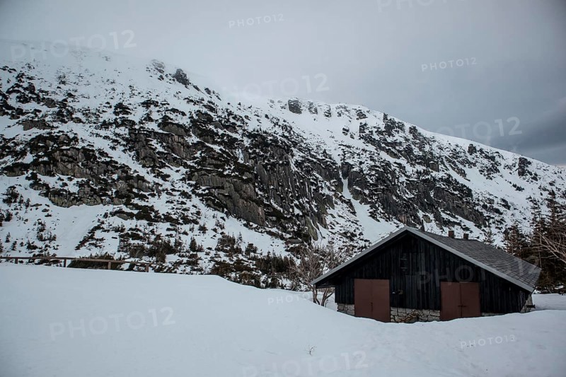 A solitary cabin sits beneath a rugged, snowy mountain landscape under an overcast sky. Poland, Karpacz, Karkonosze