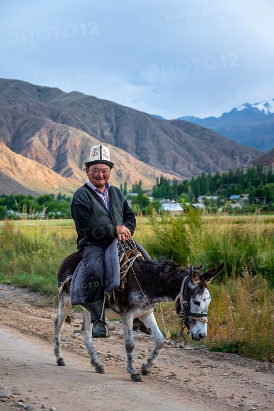 Man in traditional dress riding a donkey in front of a mountain backdrop, Kyrgyzstan, Asia