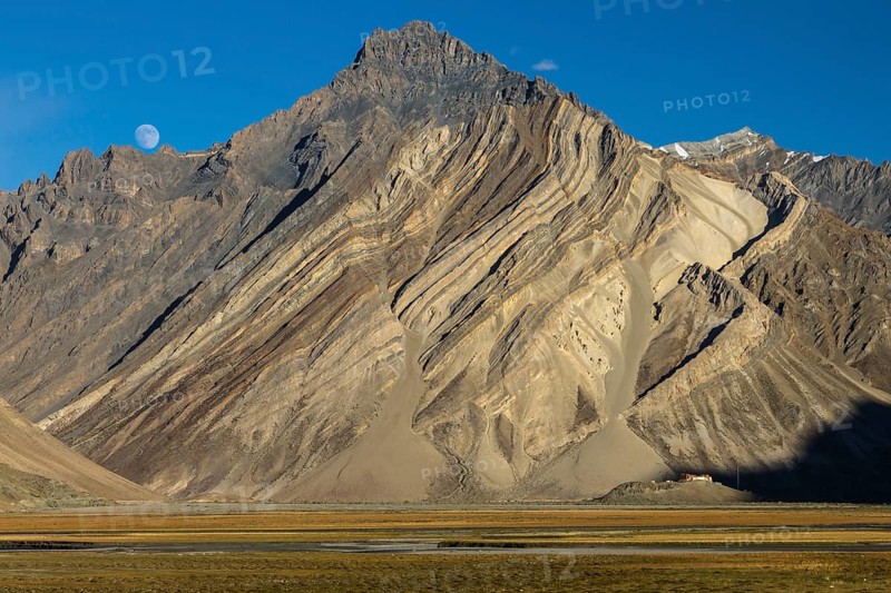 Impressive cliff in the Zanskar Mountains of the Himalayas, with clearly visible folded strata, the rock layers of sediment or metamorphic rocks, and Rangdum Gompa, the Buddhist monastery located below the rock face, on a small hill above the valley floor. Photographed in September, the late summer, at moonrise of a clear afternoon, with the moon in view in the blue sky. Kargil District, Union Territory of Ladakh, India, Asia