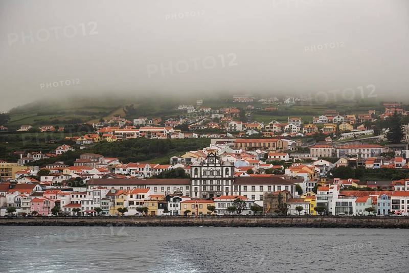The coastal town of Horta under the fog, colourful houses nestle against green hills with calm sea in the foreground, Horta, Faial, Azores, Portugal, Europe
