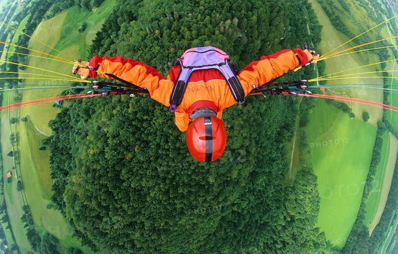 Paraglider pilot with orange clothes and red helmet flying in 500 ft above a forest and meadows, bird's eye view, Brauneck, Lenggries, Bavaria, Germany, Europe