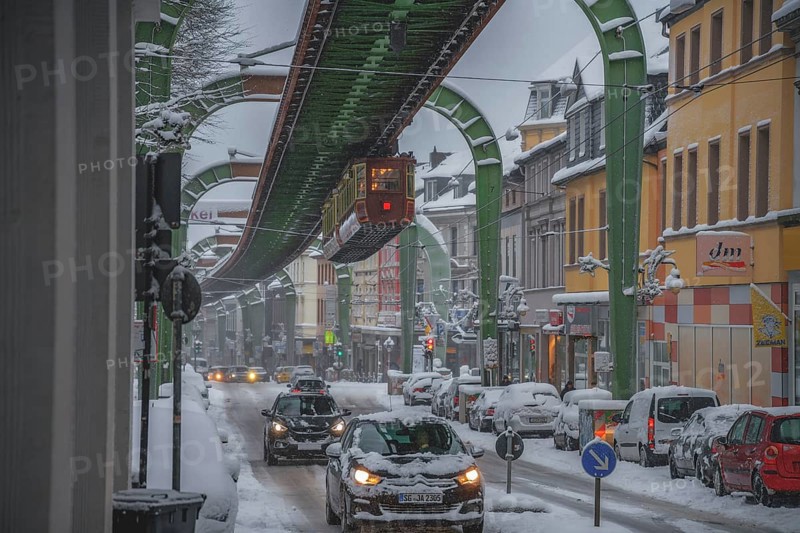 Snow-covered city street with moving cars under an overhanging railway line, wintry atmosphere, The Kaiserwagen, suspension railway, Wuppertal-Vohwinkel stop, Wuppertal Vohwinkel, North Rhine-Westphalia, Germany, Europe