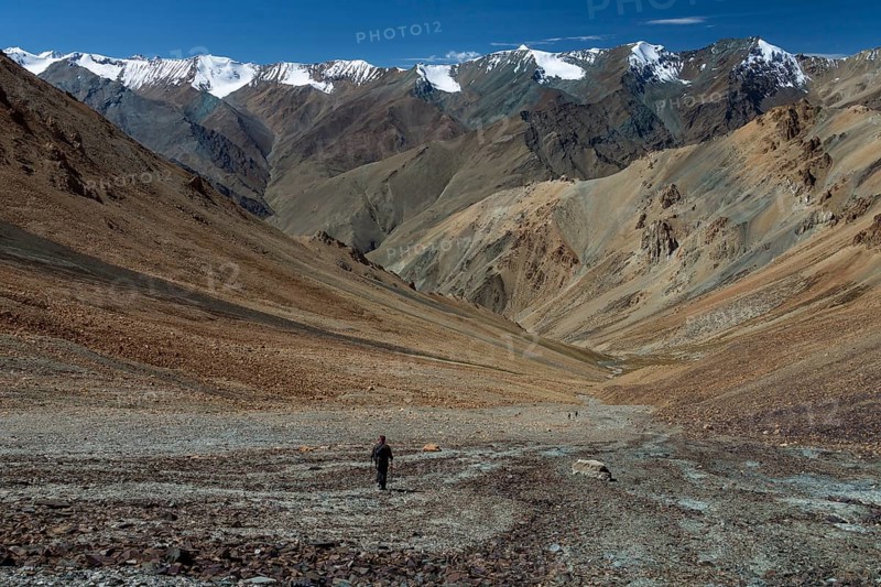 Looking down from Zherin La, a mountain pass in the remote part of the Zanskar Region, towards the Lungnak Valley, with the main ridge of the Great Himalayan Range visible on its other side. A lonely trekker is walking down the slope, and there are a few more below, in a distance. Photographed on a clear, late September day, with clear blue sky above the mountain chain. Zanskar Range of the Himalayas, dry mountains belonging to the Thetys Himalayas. Kargil District, Union Territory of Ladakh, India, Asia