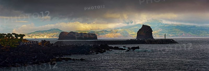 Dark clouds over a calm sea with the island of Faial in the distance and the rock formations Ilheu em Pe and Ilheu Deitado during an atmospheric sunset, Madalena, Pico, Azores, Portugal, Europe
