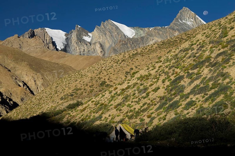 Moon setting above a trekker camp in the upper Kong Togpo Valley in Ladakh, with some high, glaciated peaks and dry slopes around, covered with scarce vegetation. Photographed late in the afternoon, on a sunny, late summer day. Zanskar Range of the Himalayas, the desert mountains belonging to the Thetys Himalayas. Kargil District, Union Territory of Ladakh, India, Asia