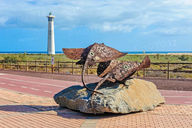 In the foreground sculpture made of scrap metal of marine animals two stingrays rays on stone pedestal, behind it developed cycle path wide cycle road with markings, left in the background lighthouse Faro de Morro Jable, Morro Jable, Jandia peninsula, Fuerteventura, Canary Islands, Canary Islands, Spain, Europe