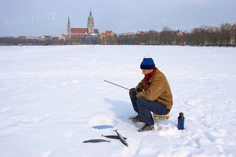 Man with winter cap or hat and warm clothes, sitting on a bucket, ice fishing on Theresienwiese at an ice hole with three mackerels lying beside him in the snow, Theresienwiese is the Oktoberfest Area, Munich, Bavaria, Germany, Europe, it is a joke, because there is no water under the snow, Europe