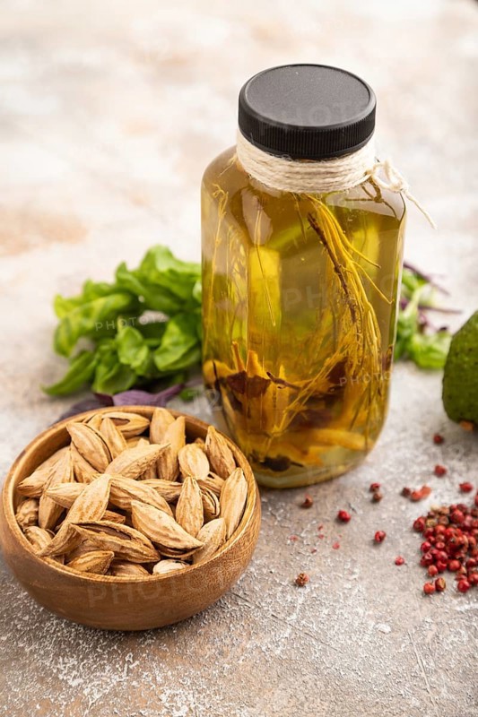 Sunflower oil in a glass jar with various herbs and spices, sesame, rosemary, avocado, basil, almond on a brown concrete background. Side view, selective focus, close up