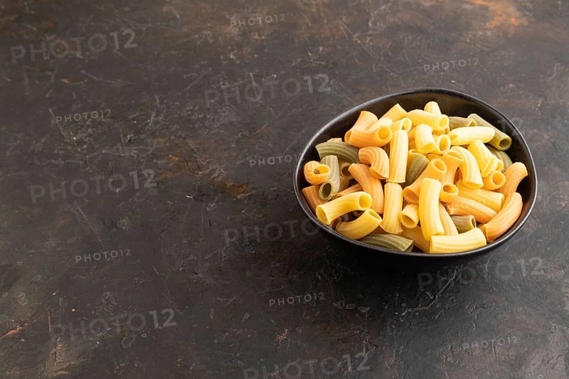 Rigatoni colored raw pasta with tomato, eggs, spices, herbs on black concrete background. Side view, copy space