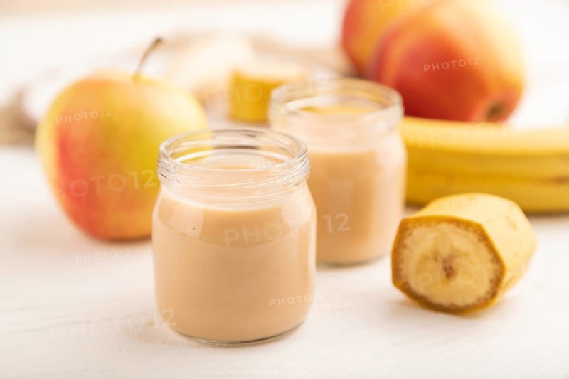 Baby puree with fruits mix, apple, banana infant formula in glass jar on white wooden background. Side view, close up, selective focus, artificial feeding concept