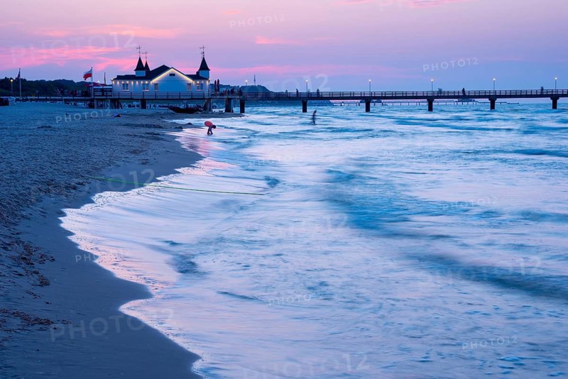 Calm evening mood on the beach with calm waves and two people near a pier with a building at dusk, clear blue-violet sky with pink clouds, Ahlbeck pier in the last evening light, motion blur, wipe effect, long exposure, view from the western beach, Ahlbeck seaside resort, Usedom Island, Mecklenburg-Western Pomerania, Germany, Europe