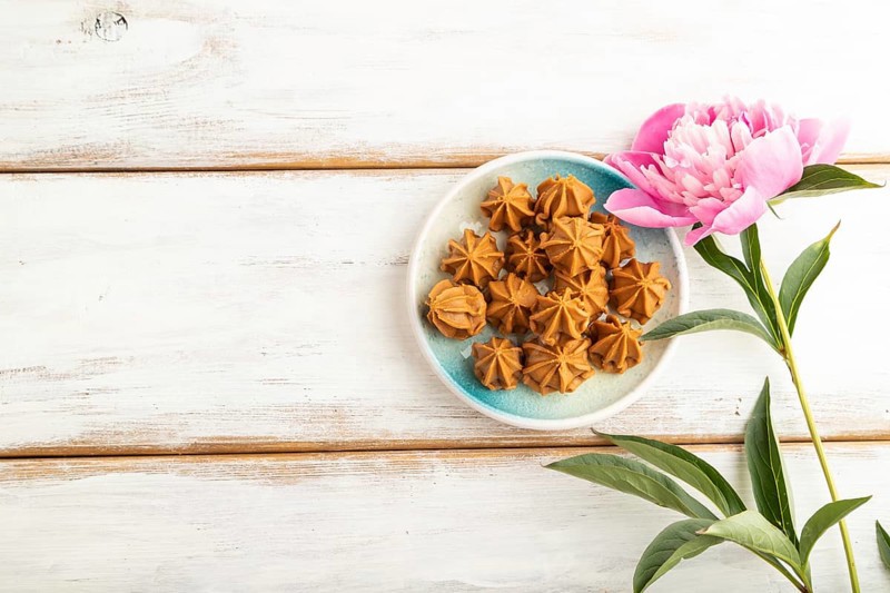 Homemade soft caramel fudge candies on blue plate on white wooden background, peony flower decoration. top view, flat lay, copy space