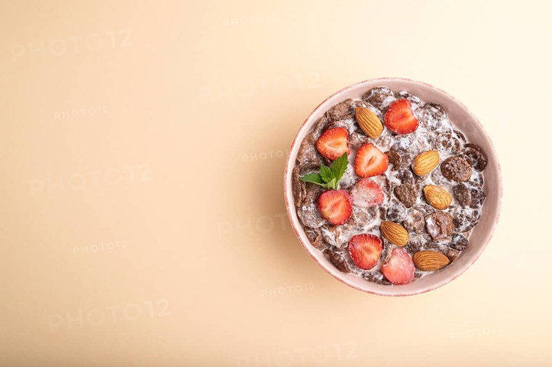 Chocolate cornflakes with milk, strawberry and almonds in ceramic bowl on pastel orange background. Top view, flat lay, copy space