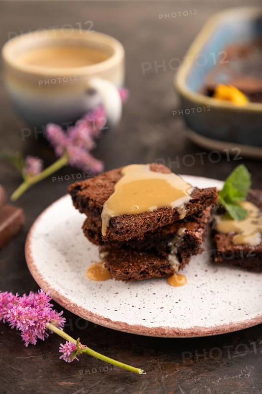 Chocolate brownie with caramel sauce with a cup of coffee on black concrete background. side view, close up, selective focus