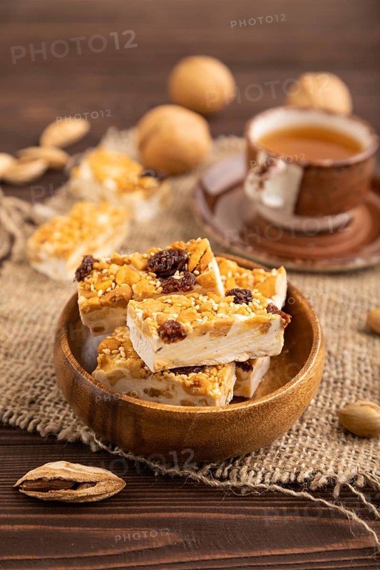 Traditional candy nougat with nuts and sesame with cup of green tea on brown wooden background and linen textile. side view, close up, selective focus