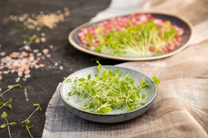 Slices of smoked cervelat salami sausage with spinach microgreen, salt and pepper on black concrete background and beige textile. Side view, close up, selective focus