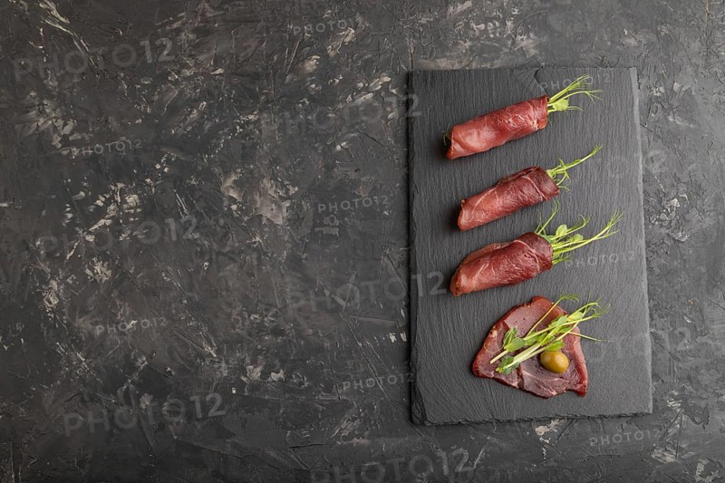 Slices of smoked salted meat with green pea microgreen on black concrete background. Top view, flat lay, copy space