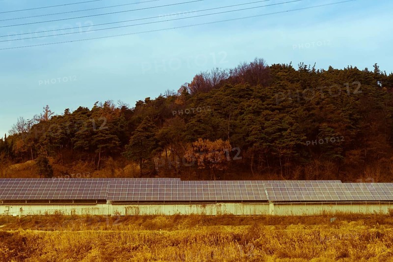 Aged photo of large array of solar panels setup in countryside in front of hillside covered in lush foliage under blue partly cloudy sky