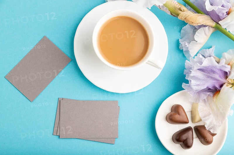 Gray business card with cup of cioffee, chocolate candies and iris flowers on blue pastel background. top view, flat lay, copy space, still life. Breakfast, morning, spring concept