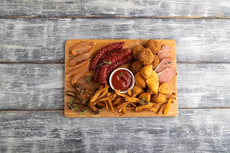 Set of snacks: sausages, fried potatoes, meat balls, dumplings, basturma on a cutting board on a gray wooden background. Top view, flat lay, close up, copy space