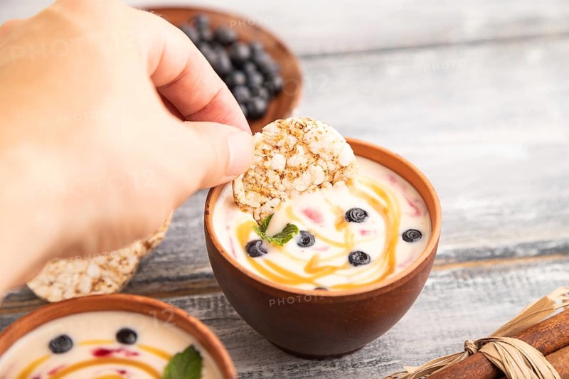 Yoghurt with bilberry and caramel in clay bowl on gray wooden, hand dipping cookies in yogurt, side view, close up, selective focus