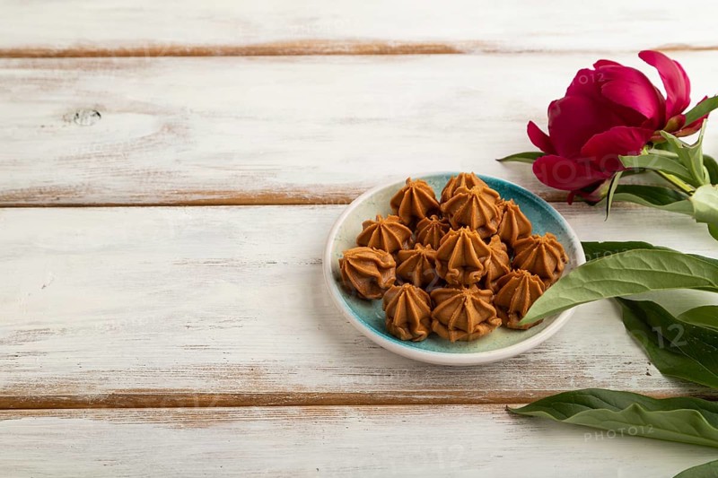 Homemade soft caramel fudge candies on blue plate on white wooden background, peony flower decoration. side view, copy space