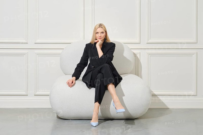 Positive businesswoman in casual suit sits on large bean bag propping chin and looking with interest