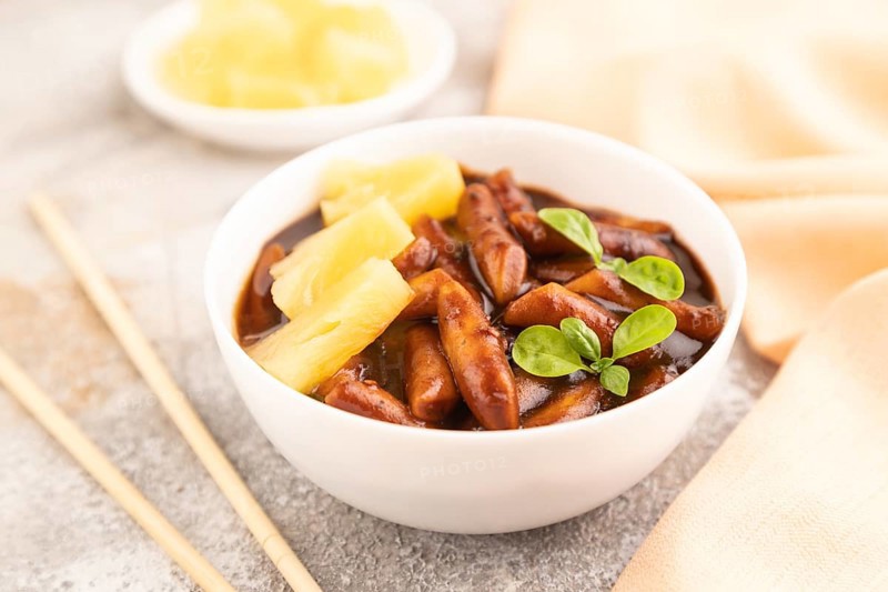 Tteokbokki or Topokki, fried rice cake stick, popular Korean street food with spicy jjajang sauce and pineapple on gray concrete background and orange textile. Side view, close up, selective focus