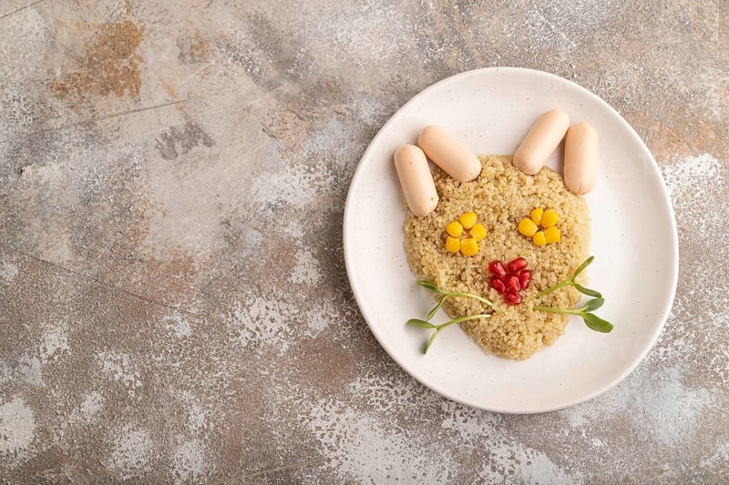 Funny mixed quinoa porridge, sweet corn, pomegranate seeds and small sausages in form of cat face on brown concrete background. Top view, flat lay, copy space. Food for children, healthy food concept