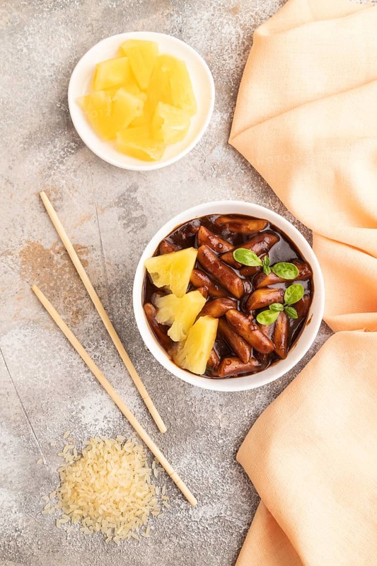 Tteokbokki or Topokki, fried rice cake stick, popular Korean street food with spicy jjajang sauce and pineapple on gray concrete background and orange textile. Top view, flat lay