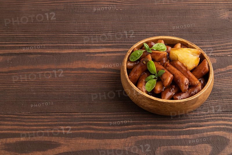 Tteokbokki or Topokki, fried rice cake stick, popular Korean street food with spicy jjajang sauce and pineapple on brown wooden background. Side view, copy space