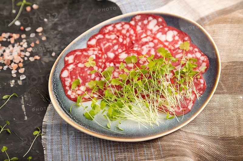 Slices of smoked cervelat salami sausage with spinach microgreen, salt and pepper on black concrete background and beige textile. Side view, close up, selective focus