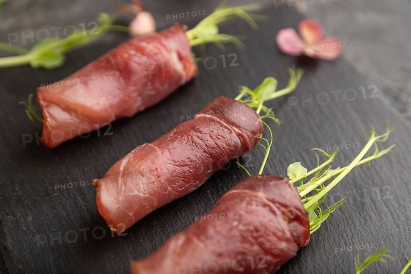 Slices of smoked salted meat with green pea microgreen on black concrete background. Side view, close up, selective focus
