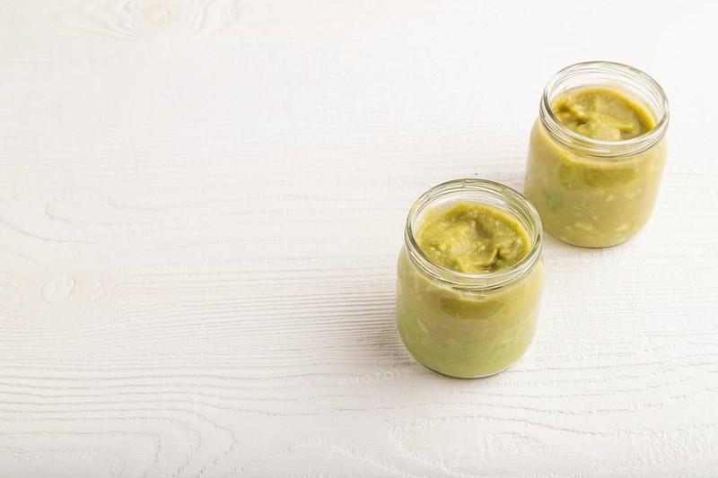 Baby puree with vegetable mix, broccoli, tomatoes, cucumber, avocado infant formula in glass jar on white wooden background. Side view, copy space, artificial feeding concept