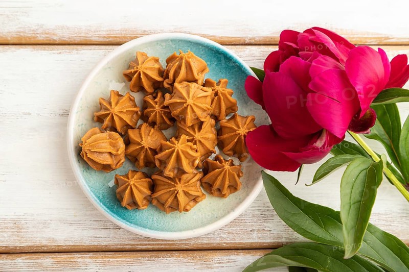 Homemade soft caramel fudge candies on blue plate on white wooden background, peony flower decoration. top view, flat lay, close up