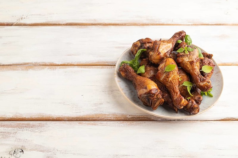 Smoked chicken legs with herbs and spices on a ceramic plate on a white wooden background. Side view, copy space