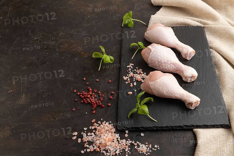 Raw chicken legs with herbs and spices on a black slate cutting board on a black concrete background and linen textile. Side view, copy space