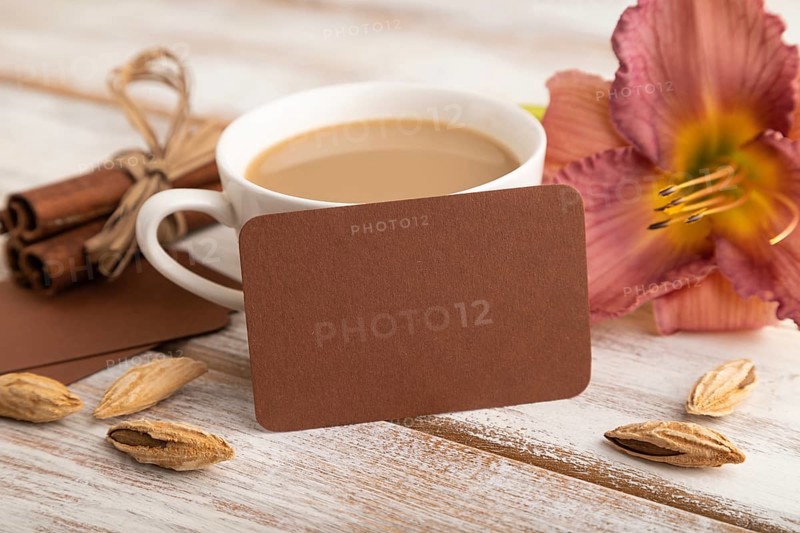 Brown paper business card mockup with purple day-lily flower and cup of coffee on white wooden background. Blank, side view, copy space, still life. spring concept