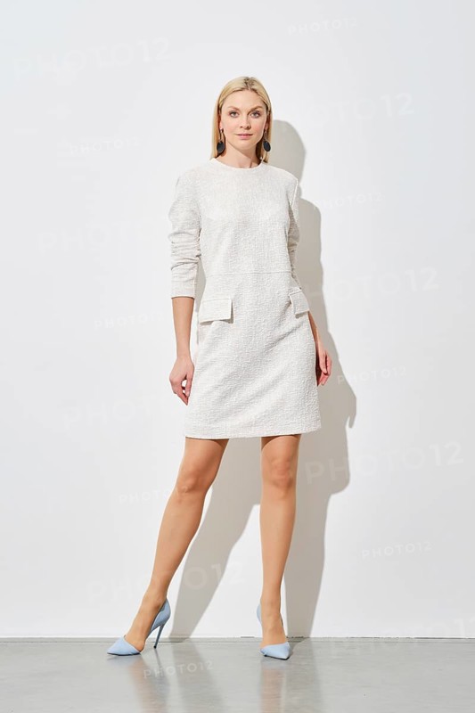 Cute woman in textured white mini dress with pockets and blue heels posing against a white wall