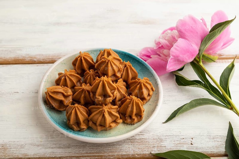 Homemade soft caramel fudge candies on blue plate on white wooden background, peony flower decoration. side view, close up