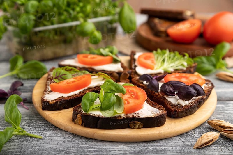 Grain rye bread sandwiches with cream cheese, tomatoes and basil microgreen on gray wooden background and linen textile. side view, close up, selective focus