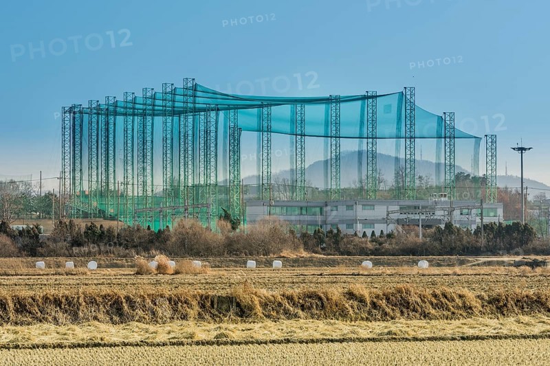 Golf driving range constructed of metal trussed columns and green netting in rural farming landscape with field of cut hay in foreground