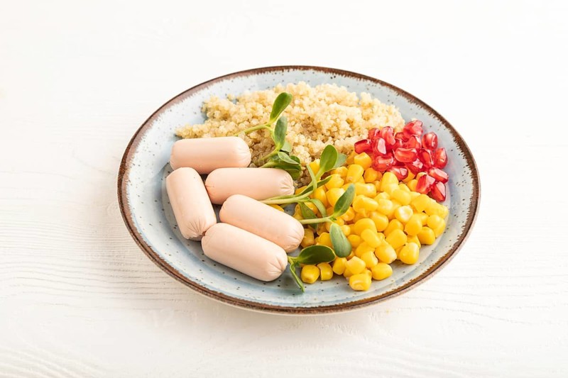 Mixed quinoa porridge, sweet corn, pomegranate seeds and small sausages on white wooden background. Side view, close up. Food for children, healthy food concept