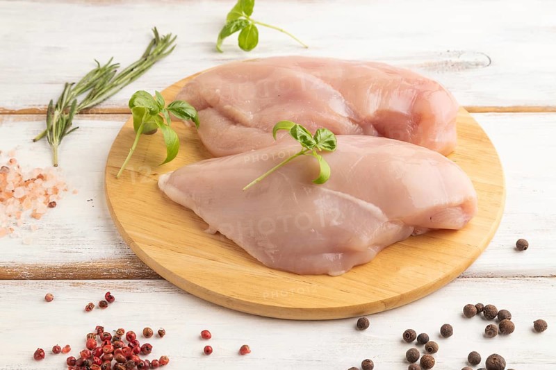 Raw chicken breast with herbs and spices on a wooden cutting board on a white wooden background. Side view, close up