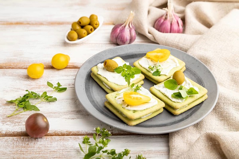 Green cracker sandwiches with cream cheese and cherry tomatoes on white wooden background and linen textile. side view, close up