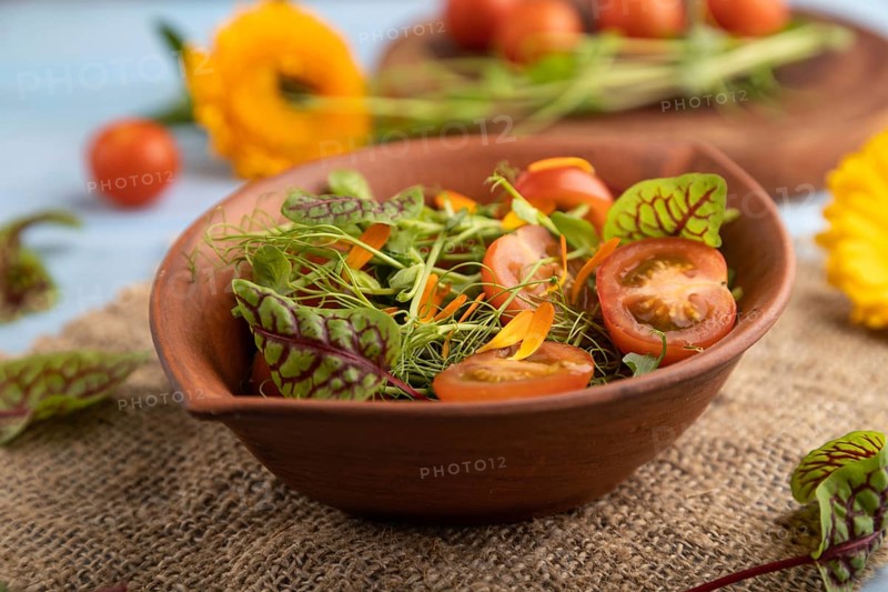 Vegetarian vegetables salad of tomatoes, marigold petals, microgreen sprouts on blue wooden background and linen textile. Side view, close up, selective focus