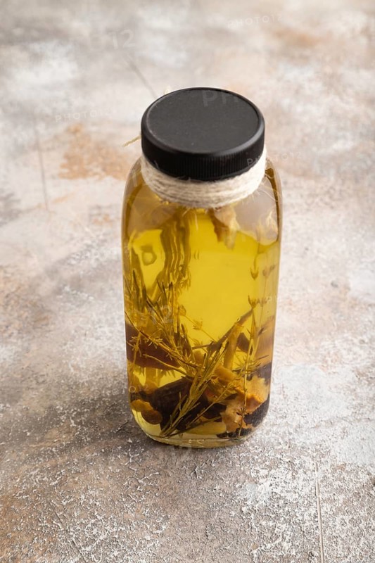 Sunflower oil in a glass jar with various herbs and spices, lavender, sesame, rosemary on a brown concrete background. Side view, close up