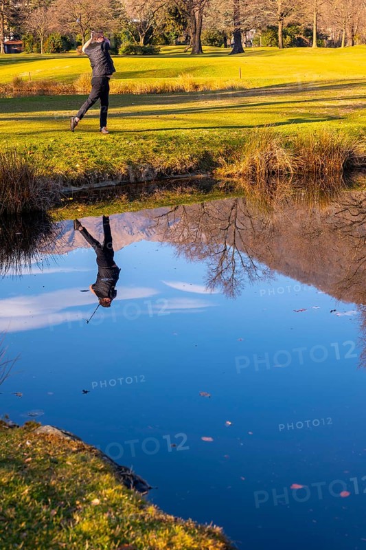 Male Golfer Reflected in a Water Pond and Hitting the Golf Ball on Fairway on Golf Course with Mountain in a Sunny Day in Switzerland