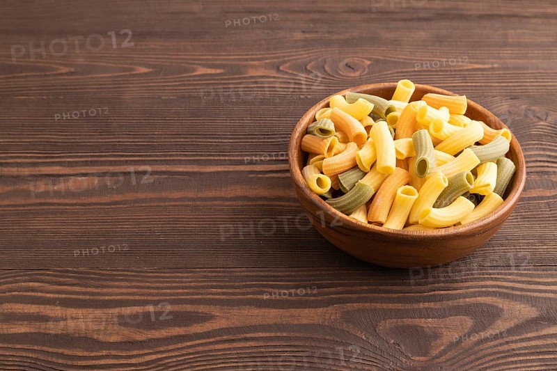 Rigatoni colored raw pasta with tomato, eggs, spices, herbs on brown wooden background. Side view, copy space